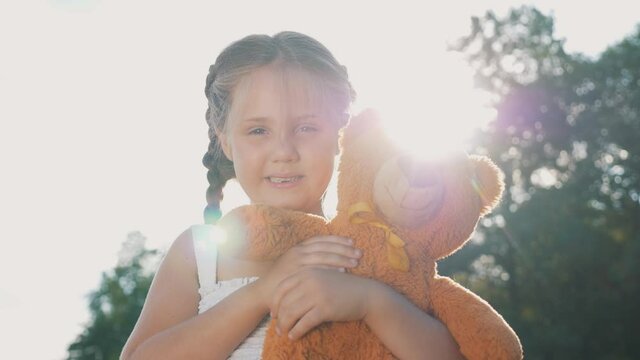 Little girl with a toy teddy bear in park. Cute girl with a teddy bear in a green field. Lonely girl with a toy teddy bear in park in green field.Teddy bear in the hands of a girl in the park