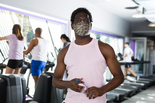 Positive man wearing face mask for viral protection resting after exercises in fitness gym holding drinking water