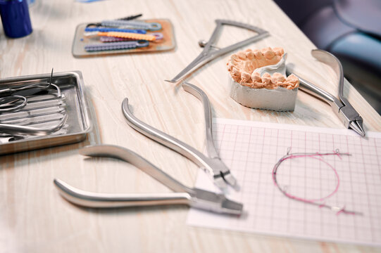 Desk with orthodontic tools, ligature ties, dental teeth model and wire braces drawing. Ligature wire cutters, pliers, multicolored teeth correctors and lower jaw model on table in dental office.