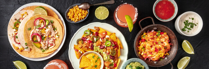 Mexican food, a flat lay panorama on a dark background. Nachos, tacos, Paloma cocktails, and various dips