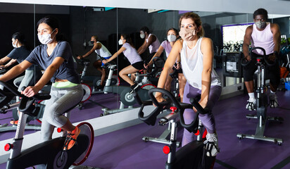 Fototapeta na wymiar Multiethnic group of young adults in protective face masks training on cycling machines in gym during coronavirus pandemic