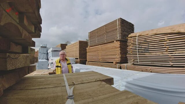 Pensioner in glasses and yellow warm vest walks about open industrial wood storehouse located in port area against cloudy sky