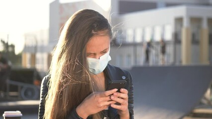 young girl in protective medical mask walks down street, with phone in hand. girl reads news of the covid 19 treatment. coronavirus, pandemic virus protection mask. girl walks around city with phone. - Powered by Adobe