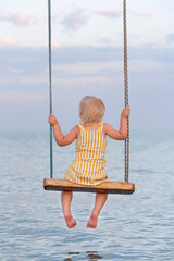 Toddler sitting on rope swing over the water. Back view. Vertical frame. Happy childhood.