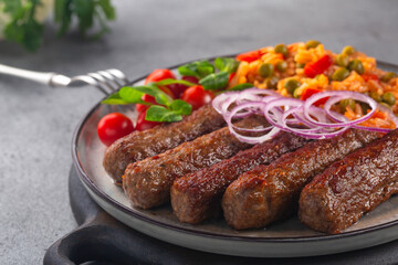Close-up of beef kofta or cevapcici with rice and vegetables, decorated with red onion. On grey table.