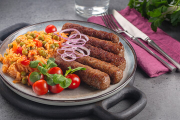 Balkan dinner with beef cevapcici and djuvec rice and vegetables, decorated with red onion. On grey table. - obrazy, fototapety, plakaty