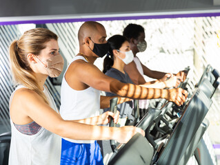 Active people in protective masks having running elliptical trainer class in health club