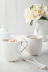Two white porcelain cups of coffee. Bouquet of delicate cream flowers in the background. Romantic morning for two.