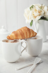 Two white porcelain cups of coffee with croissant. Bouquet of delicate cream flowers in the background. Romantic morning for two