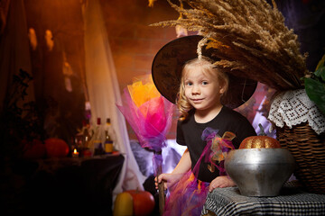 Little cute blonde girl looking as witch in special dress and hat in room decorated for Halloween....