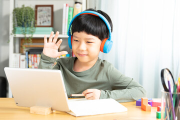 Young happy smart looking Asian student boy video call and wave his hand to computer tablet in online learning class. Study at home, New normal in technology and education during Covid-19 lockdown