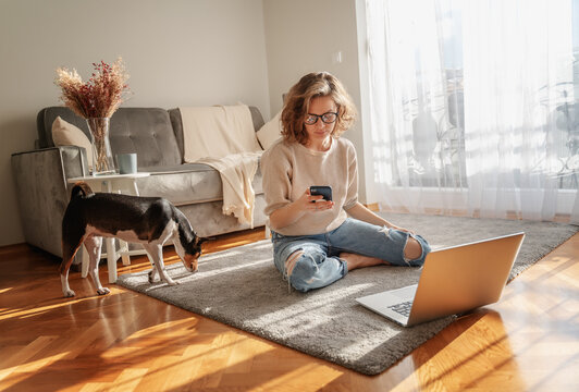 Pretty Curly Happy Young Woman Sitting At Home On The Floor In Front Of Laptop With Her Pet Dog Working And Learning Online With Smartphone In Hands