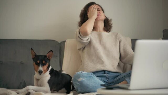 A woman having a migraine attack while works on a laptop while sitting on the couch with her dog in the living room