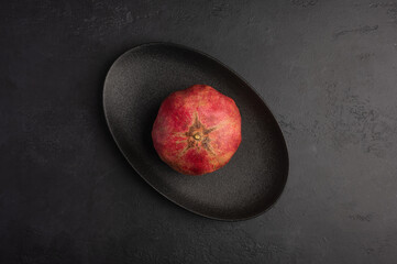 One pomegranate in the peel served in black oval plate on moody dark background. Top view, healthy...