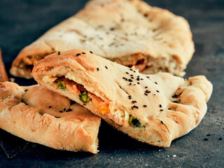 Perfect homemade closed pizza calzone on baking paper sheets over black background. Cutting tasty italian closed calzone pizza with cheese, meat, vegetables, black sesame dressed. Copy space for text