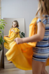 A little girl tries on her mother's yellow dress in front of the mirror. Children imitate adults
