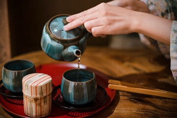 Fototapeta na wymiar woman hand Pouring green tea into cup. Traditional japanese chinese iron teapot and ceramic teacups, asian vintage style tea set on wooden table. 