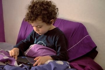 4-5 year old boy with curly hair lying down watching cartoons on tablet, Happy boy lying in bed...