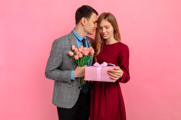 Handsome elegant man in classic suit giving flowers and gift box to his beautiful girlfriend on pink background, Valentine's Day concept