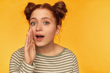 Teenage girl, happy looking red hair woman with two buns. Wearing striped blouse and holding hand next to her mouth, whisper to you. Watching at the camera isolated over yellow background