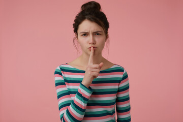 Young lady, pretty woman with brunette hair and bun. Wearing striped blouse and showing sign of silence, touching lips with a finger. Watching at the camera isolated over pastel pink background