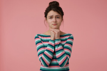 Portrait of asking, sad looking girl with brunette hair and bun. Wearing striped blouse and hold hands under her chin. Emotional concept. Watching at the camera isolated over pastel pink background