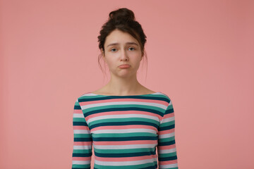 Young lady, unhappy woman with brunette hair and bun. Wearing striped blouse and pout a lip, offended. Emotional concept. Watching at the camera isolated over pastel pink background