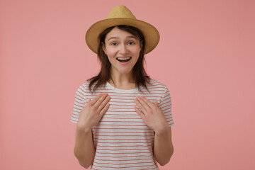 Portrait of attractive, adult girl with long brunette hair. Wearing t-shirt with red strips and hat. Pointing at her self. Emotion concept. Watching at the camera isolated over pastel pink background