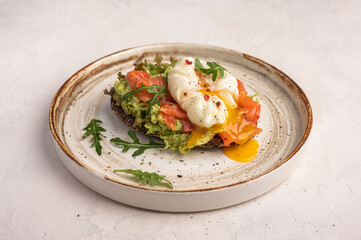 Homemade poached egg toast with avocado, salted salmon, arugula and rye bread on a ceramic plate, close up. Healthy food concept