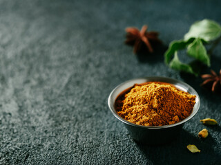 Indian or pakistani masala powder and spices on dark textured background. Small metall bowl with dry curry garam masala mix spices blend, green bay leaf, cardamom and star anise with copy space left