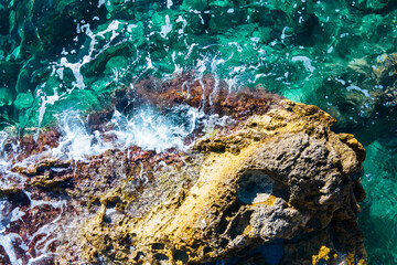 Rock and reef covered with moss and algae washed by foam waves in the sea or ocean with clear water on nice sunny day, top view from height, drone shooting.