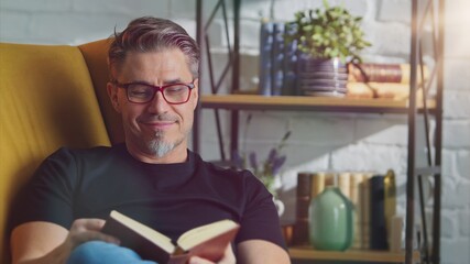 Older white man in glasses reading book at home sitting in armchair in cosy room.