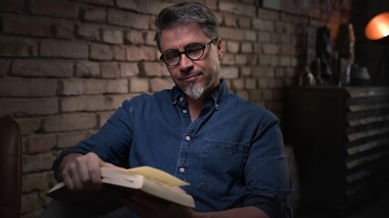 Mature man in his 50s reading book at home sitting in dark living room.