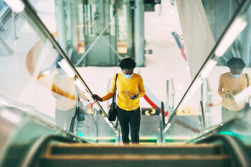 A young African woman in a protective medical mask climbs the escalator in the subway and listens to music with headphones. Modern technologies, pandemic, social distance