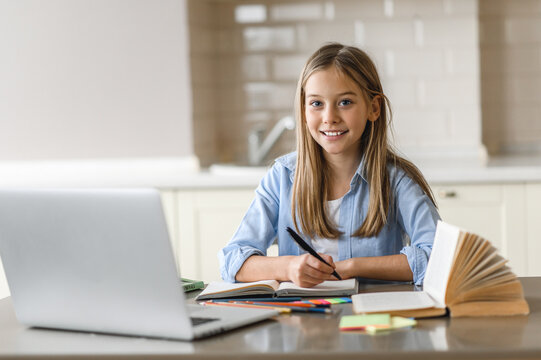 Caucasian Pretty Blonde Schoolgirl Is Learning At Home Using Laptop, Sitting At The Work Desk, Looks At The Camera And Smiling. Education During Quarantine