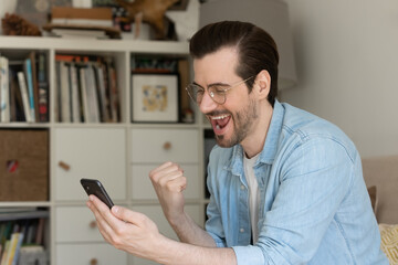 Overjoyed with good news. Laughing young man read email message from phone screen scream yes show...
