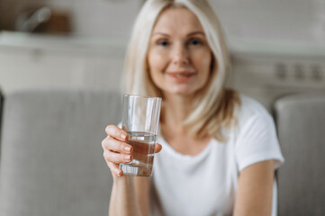 Healthy lifestyle concept. Smiling caucasian blonde female mature in out of focus sits on the sofa in living room, holding a glass of water in a hand in the foreground, follow healthy lifestyle