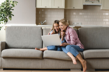 Fototapeta na wymiar Mom and daughter spend time together at home. Happy caucasian mom and daughter are sitting on the couch, using a laptop, laughing, browsing the Internet, shopping online, seeing the good news