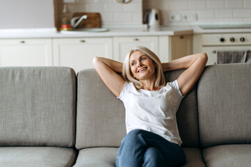 Happy mature caucasian woman is relaxing on the couch in living room at home. Middle aged woman enjoying weekend or leisure from homework sitting on sofa, looks away and smiling