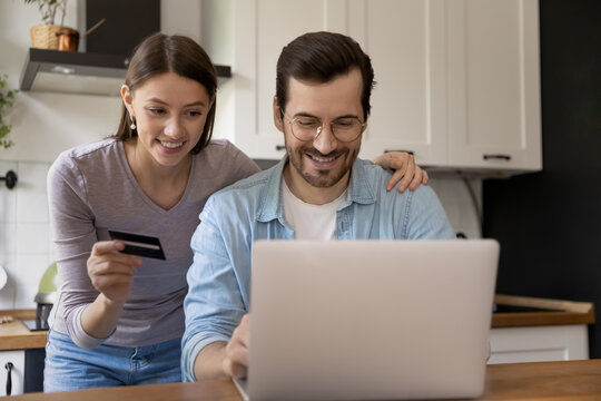 Visiting online shop. Happy young spouses engaged in internet shopping by laptop computer at home kitchen. Husband and wife ordering buying consumer goods at web store making payment by credit card