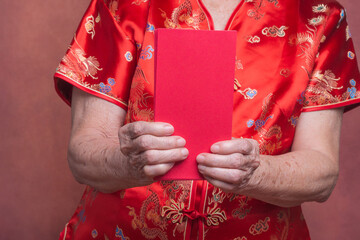 Hands of a senior woman holding red envelopes (Angpao) and wearing a traditional cheongsam qipao dress while standing in the studio with a vintage background. Close-up photo. Chinese New Year