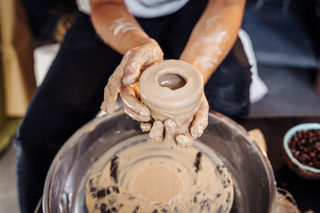 Close-up of the hands of a woman ceramist working with a potter's wheel in a cozy, bright workshop. A young experienced woman skillfully makes a mug, vase or plate. Creative people, pottery workshop