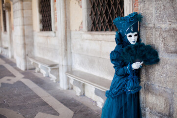 Fototapeta na wymiar Masked woman in ornate costume at the the Venetian masquerade stands against stone wall near St. Mark's Square in Venice