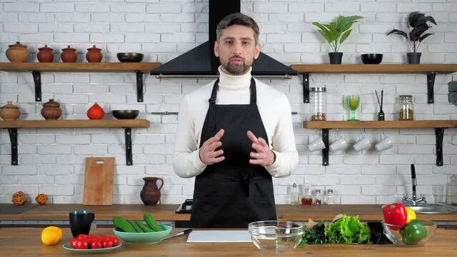 Confident smiling man chef in black apron looking camera tells teaches records remote online video culinary webinar master class course in home kitchen, vegetables ingredients cooking salad on table