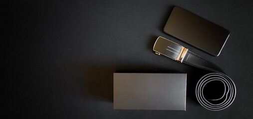 Black leather belt with golden automatic buckle, smartphone and gift box lie on a dark background. Presents for man. Free space for an inscription.