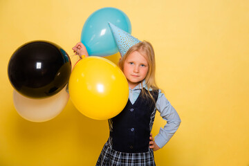Obraz premium A girl in a school uniform with a festive cap on her head is holding balloons in her hands isolated on a yellow background.