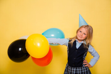 Fototapeta premium A girl in a school uniform with a festive cap on her head is holding balloons in her hands isolated on a yellow background.