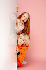 Astonished. Happy children isolated on coral pink studio background. Look happy, cheerful. Copyspace for ad. Childhood, education, emotions, facial expression concept. Peeking out from behind the wall