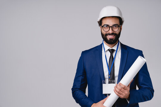 Cheerful young Arabian engineer with architectural plan and hardhat isolated over grey background