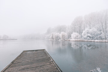 Frozen lake in winter, with wooden pier, and trees covered by snow, beautiful foggy sunset on the forest. Beautiful seasonal winter landscape, peaceful nature scenery